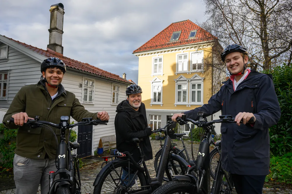 Three smiling people with helmets and black e-bikes on a cobbled street between white and yellow wooden houses