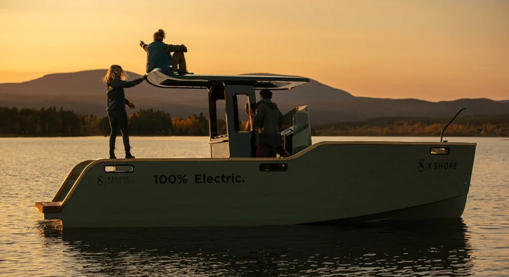 The X Shore electric boat on a calm fjord at sunset, two people sitting on its roof