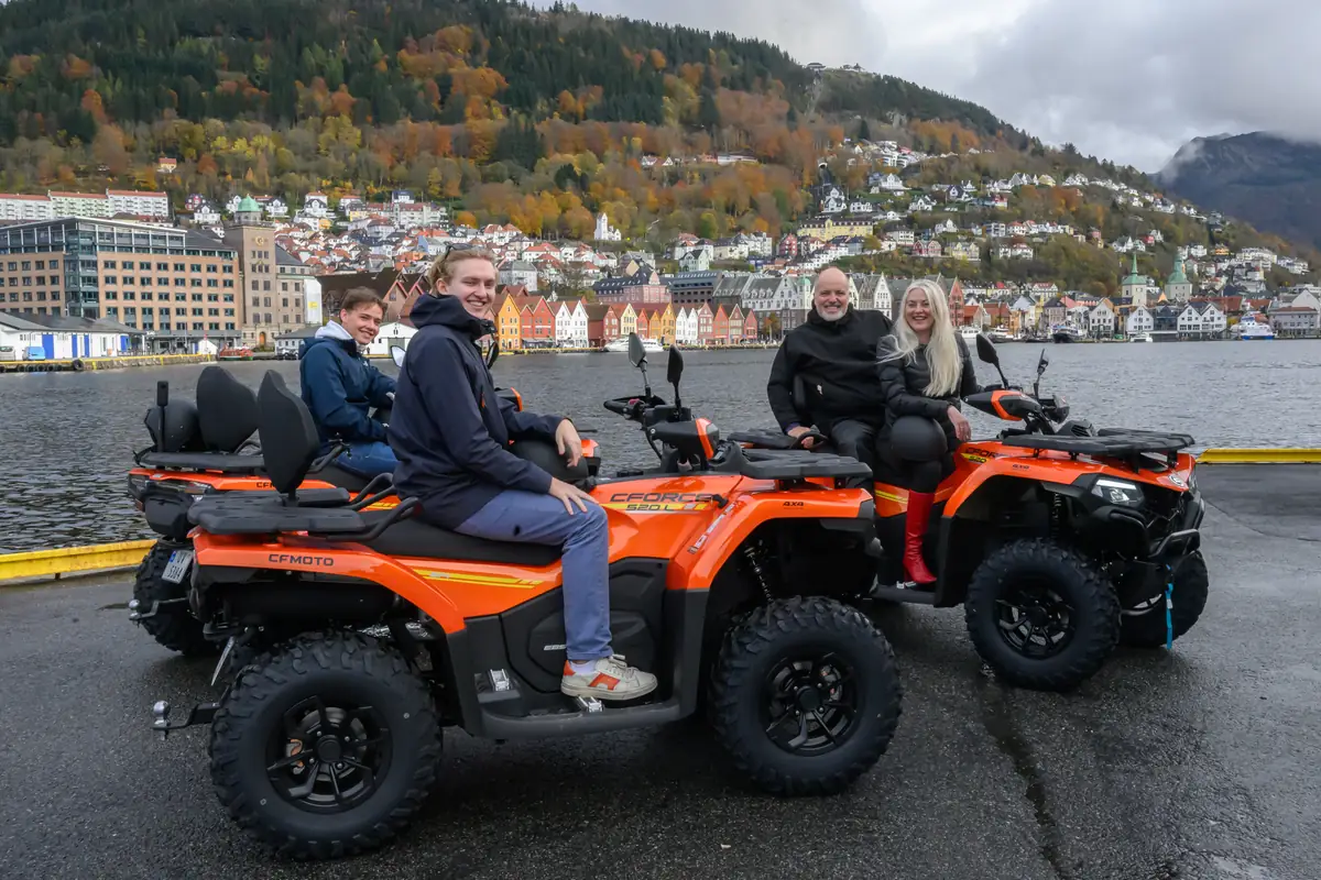 Four people on two orange quad bikes on the quayside, with the wooden houses of Bryggen and the autumn hillside behind them