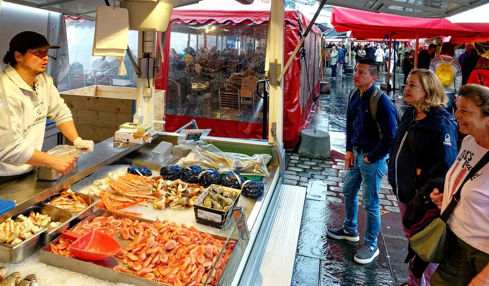 An outdoor stall at Bergen's fish market, king crab legs and prawns on ice, visitors looking on in the rain