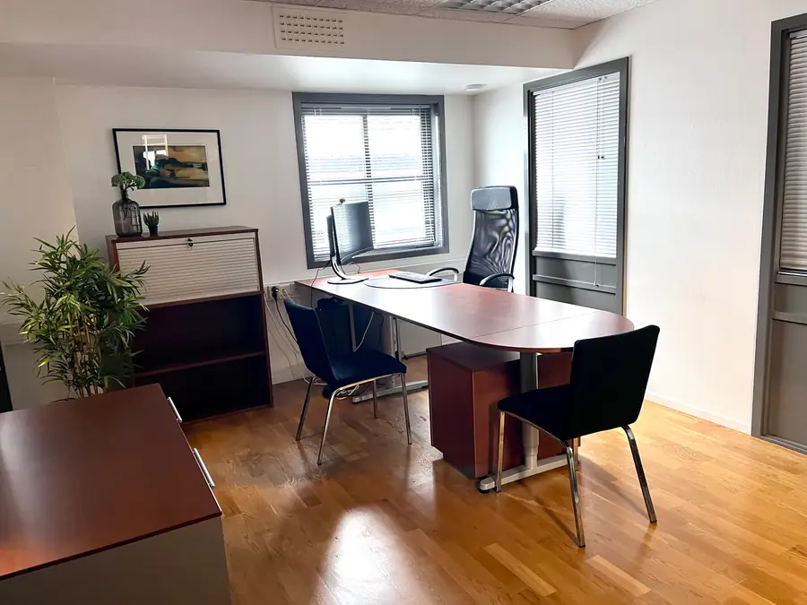 Kontor med skrivebord, skjerm og to vinduer med persienner