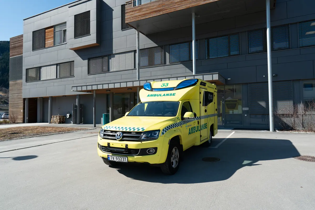 Gul norsk ambulanse parkert utenfor hovedinngangen til et moderne helsesenter