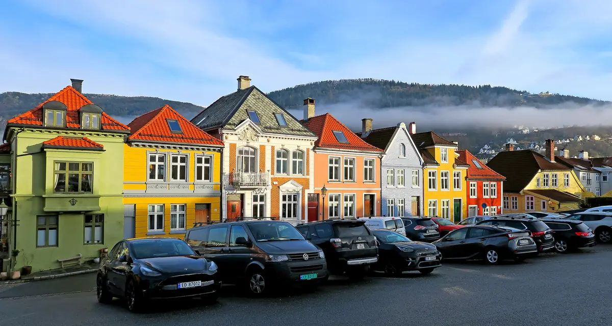 Rekke med fargede trehus på Klosteret i Bergen, med parkerte biler foran og skogkledd fjellside i tåke bak