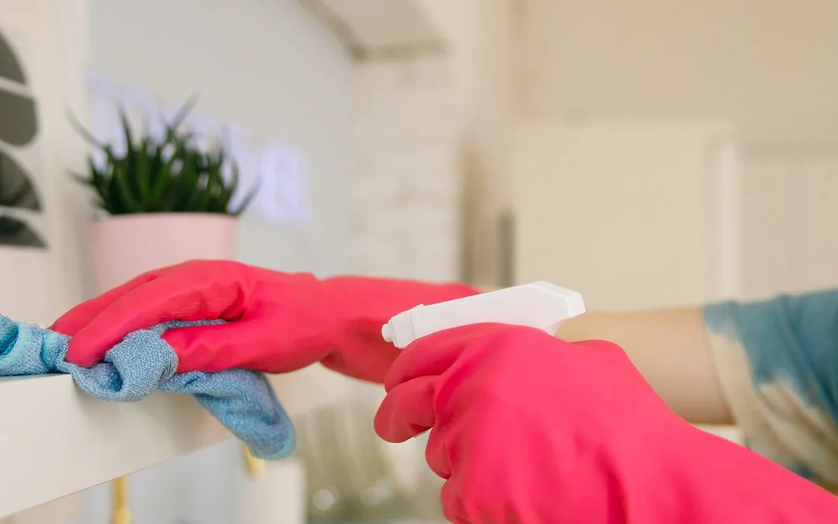 To hender i rosa gummihansker tørker av en hylle med klut og sprayflaske